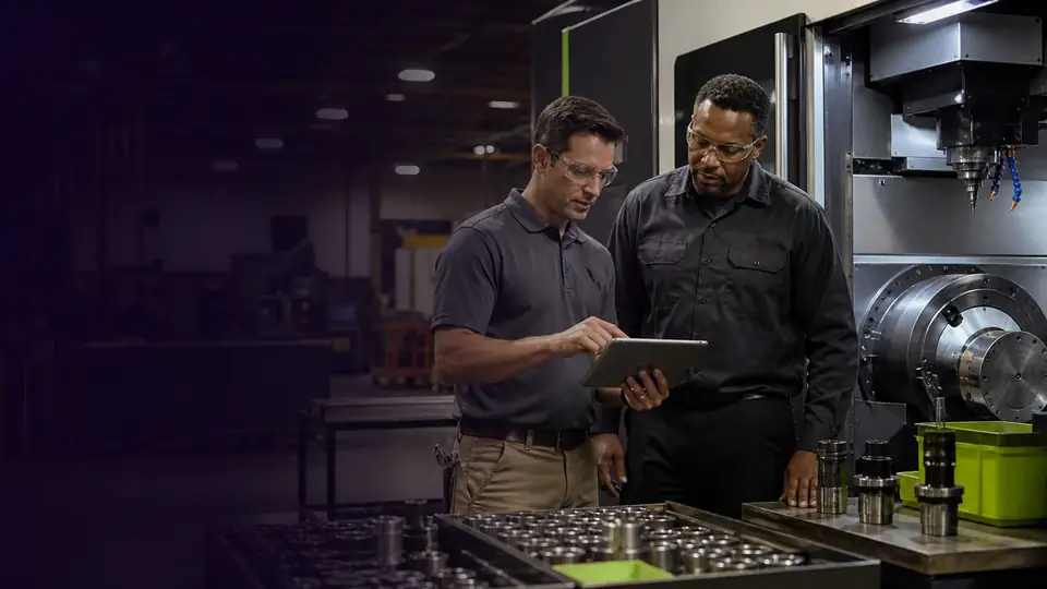 Two machinists reviewing specifications on a tablet beside a CNC machine.