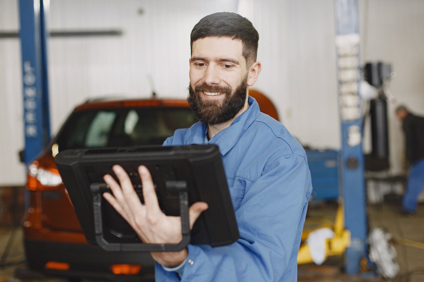 Smiling mechanic holding a tablet in an auto repair shop with a car on the lift behind him