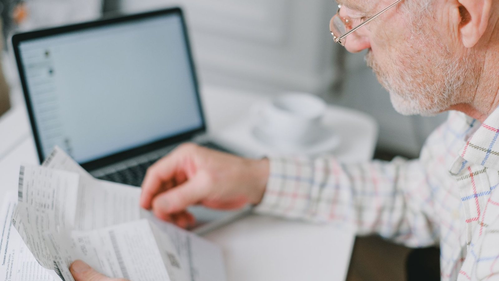Man wearing glasses reading a printed document beside an open laptop