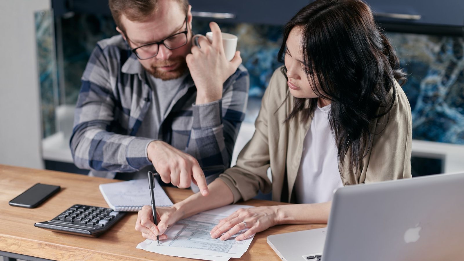 Two business owners reviewing costs on paper with a calculator and laptop at a kitchen table