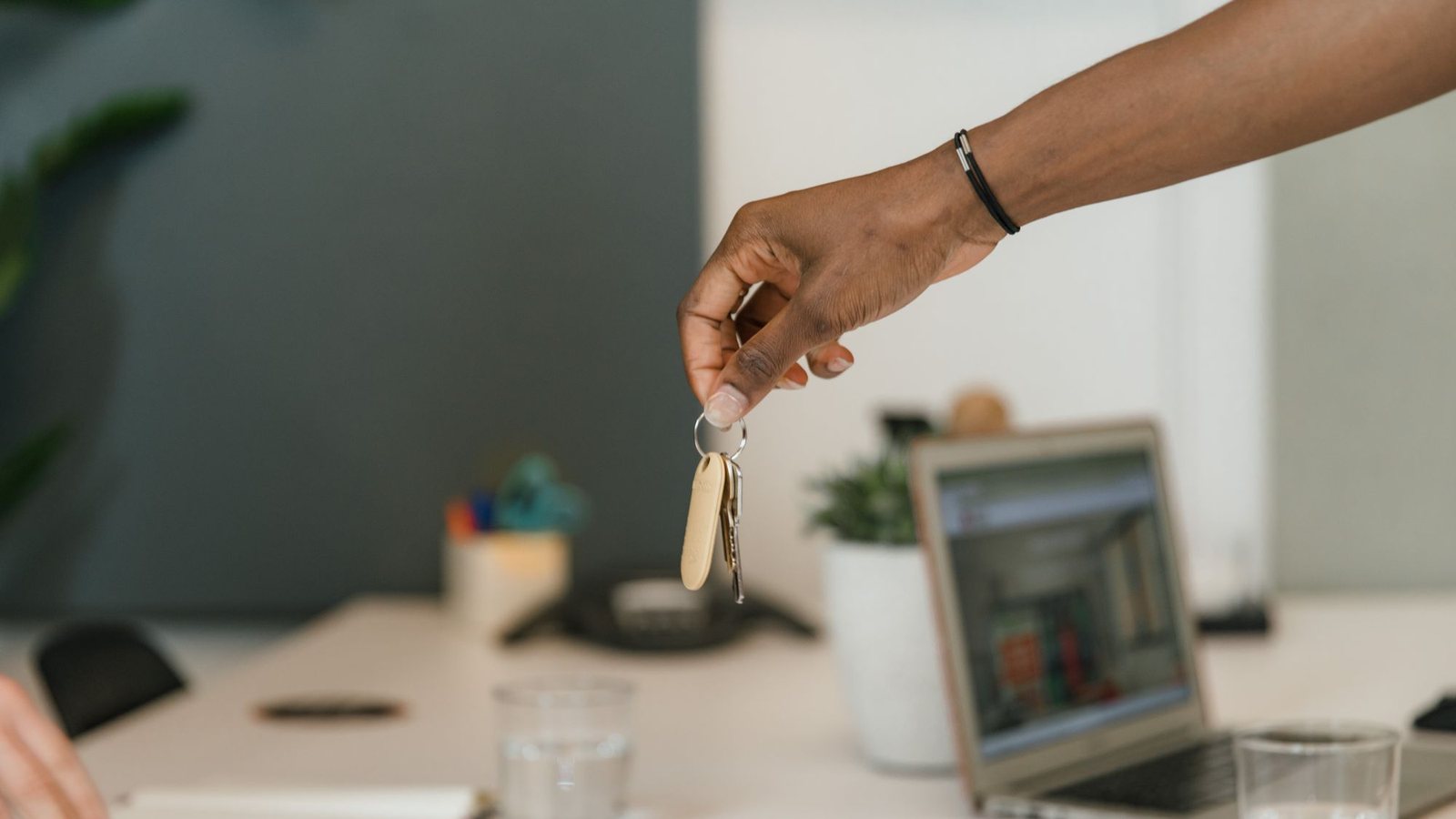 Hand passing a set of keys across an office desk with an open laptop in the background