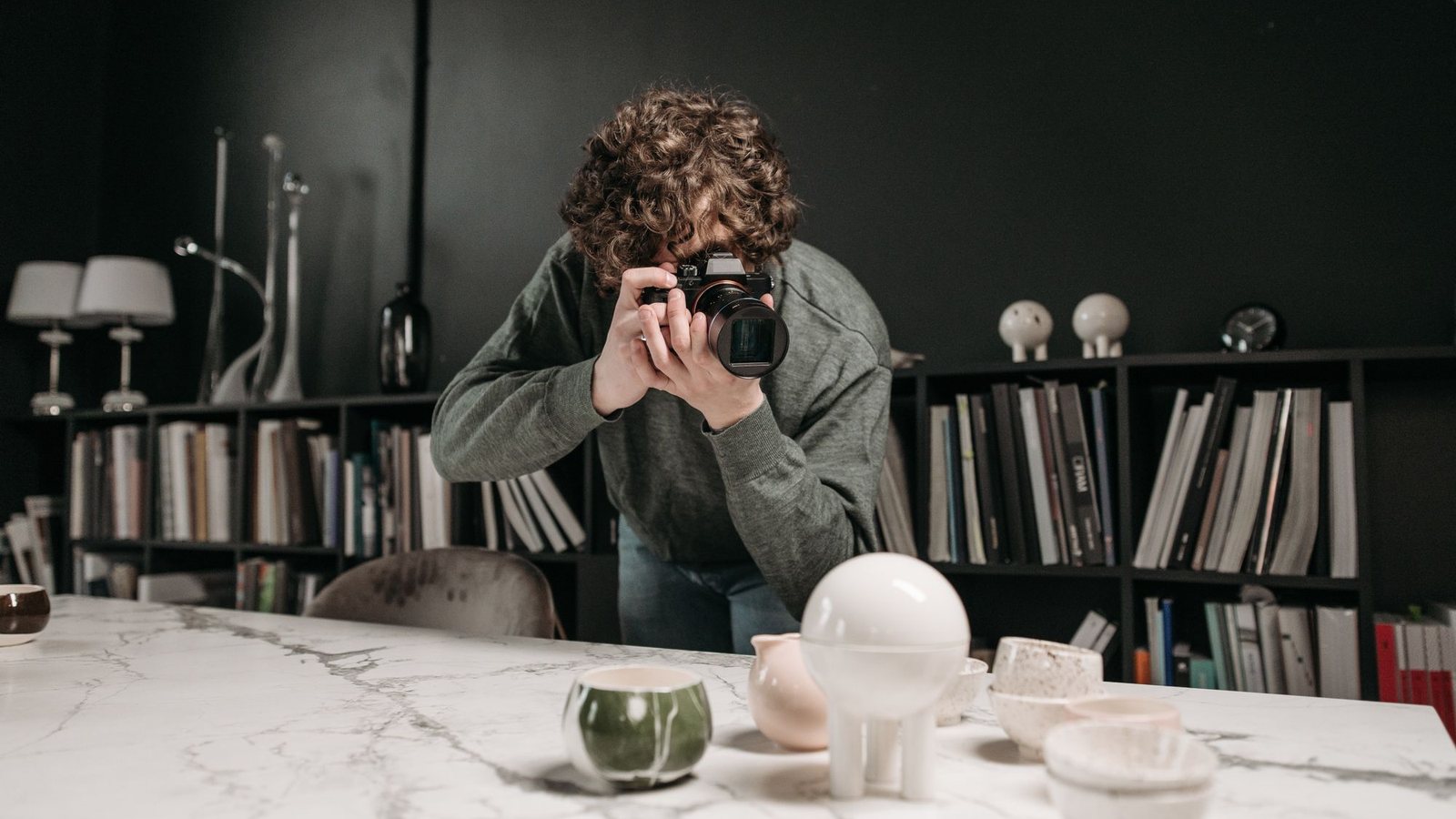 Photographer taking pictures of cups and vases on a marble table, with bookshelves behind